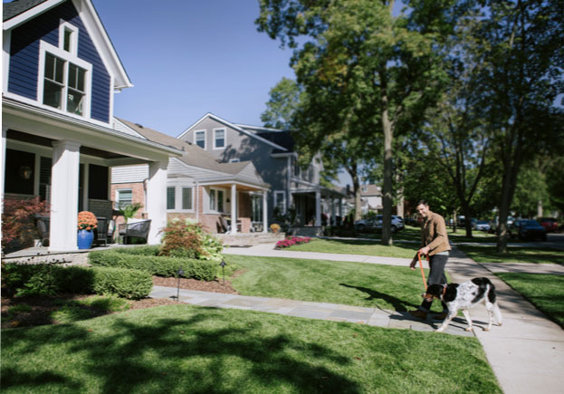 A man walks his dog up the sidewalk toward a craftsman-style home.