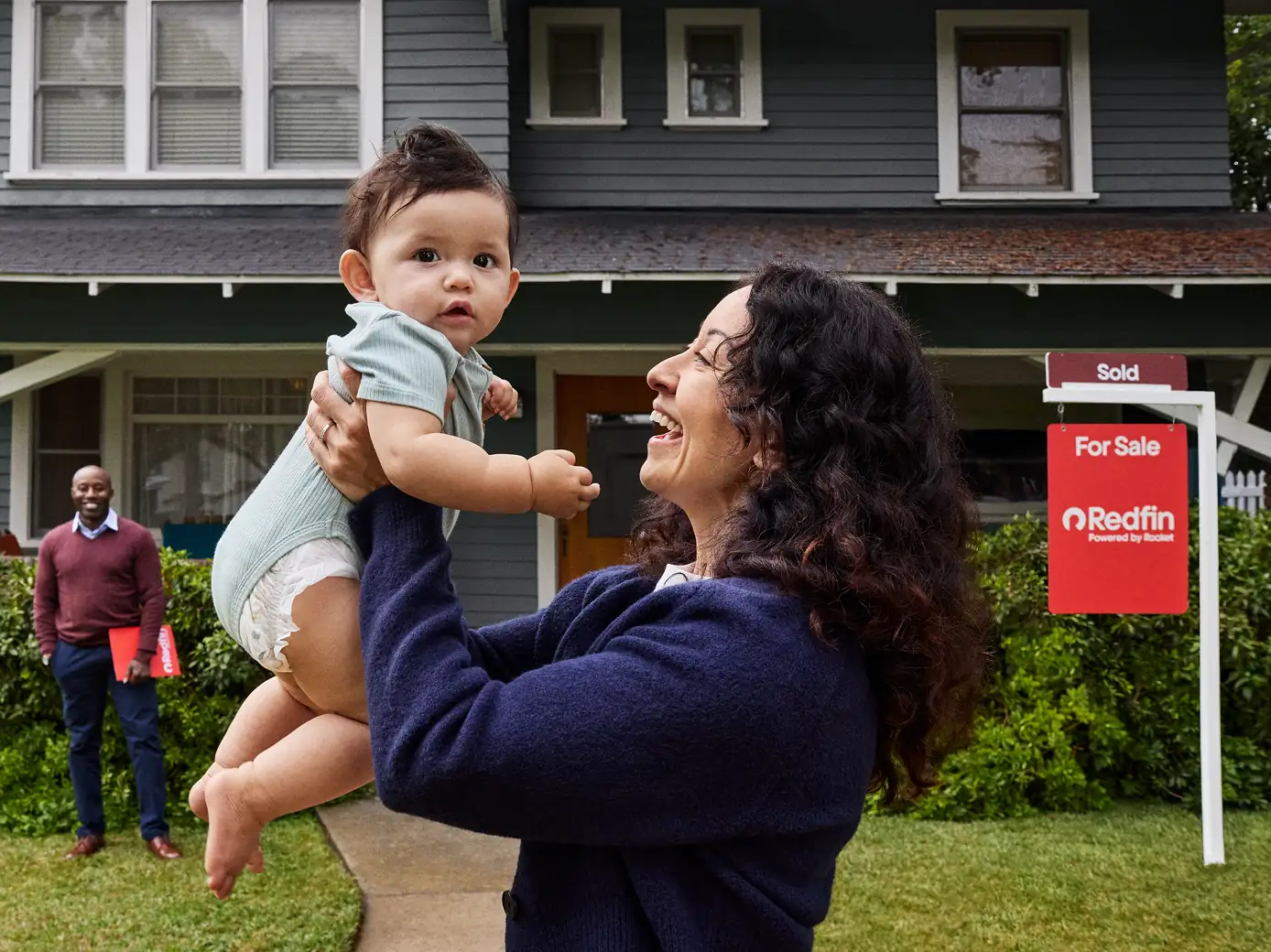 A woman lifting a baby in front of a green two-story house with a Redfin “Sold” sign in the yard while the realtor stands behind them. A tag says Bundle and save.