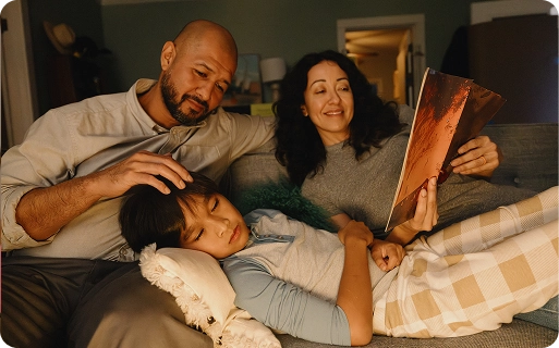Two parents sitting on a couch with a child resting on a pillow, while the mother reads a book.