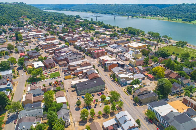 Aerial view of Madison Indiana along the Ohio River with blue bridge, hills and many trees in the background.