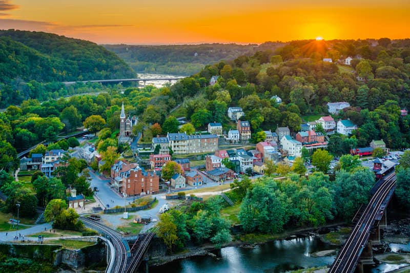 View of Harpers Ferry at sunset with lush trees, river and downtown area.