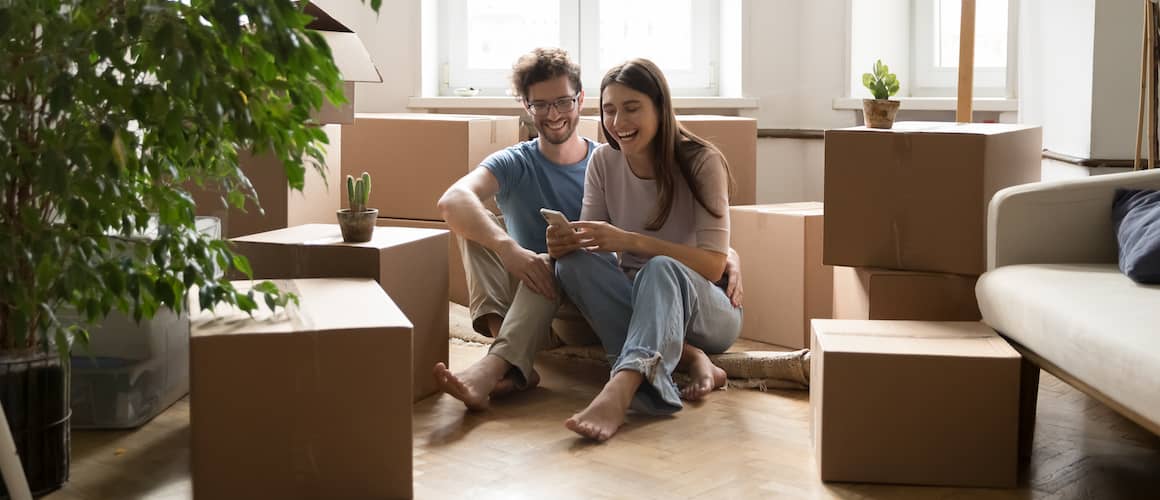 Millennial couple sitting on floor of new home, surrounded by moving boxes and houseplants.