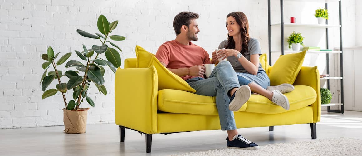 Young Couple sitting together on a yellow sofa talking and drinking out of mugs.