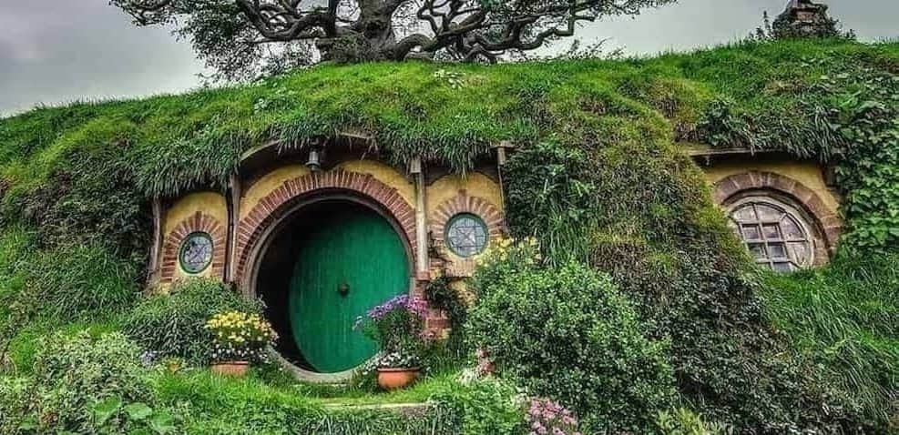 Hillside home covered with leafy vegetation and featuring a circular door and windows. Home is photographed against a cloudy sky with a large tree in the background.