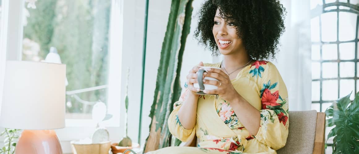 Lady wearing yellow dress sitting near lamp drinking coffee.