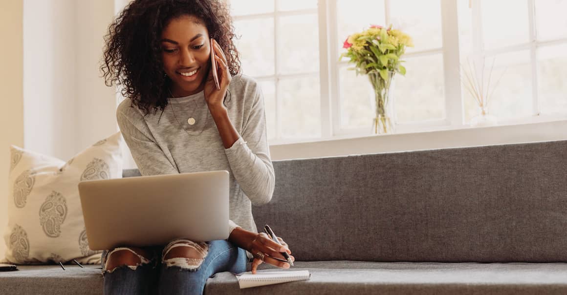 Woman sitting on couch looking at laptop while talking on phone.