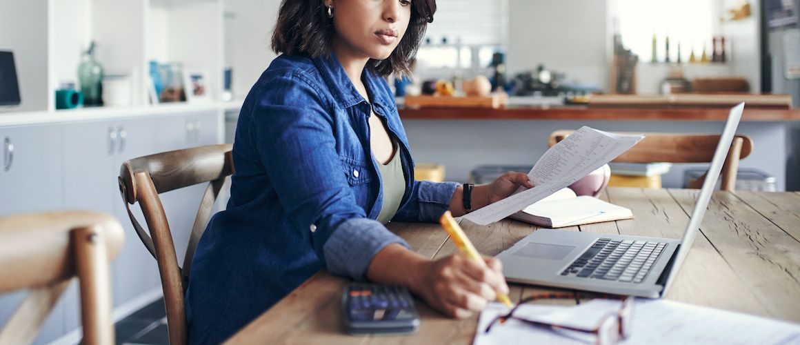 Woman looking over her finances as her kitchen table at home, with a laptop in front of her and paper.