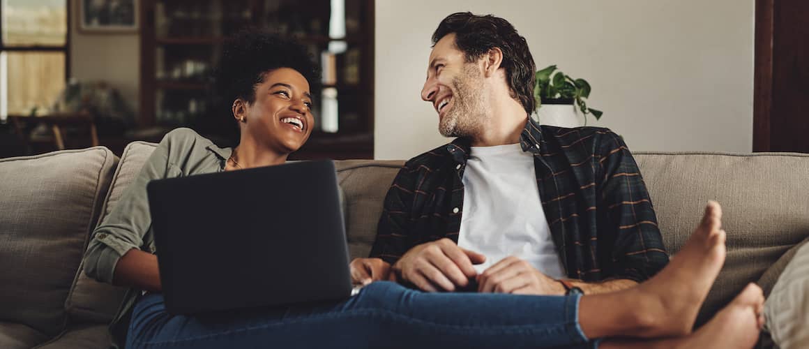 Happy couple on couch looking at each other and holding a laptop.