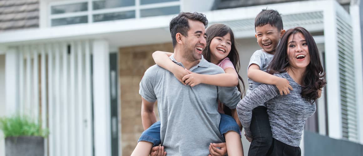 Couple With Two Kids Giving PiggyBack Rides In Front Of House