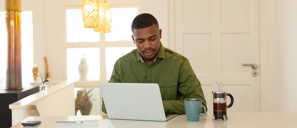 Man In Green Sitting At Counter With French Press And Laptop
