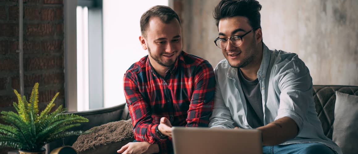 Same-Sex Male Couple Spend Time Together Drinking Coffee And Looking At Laptop