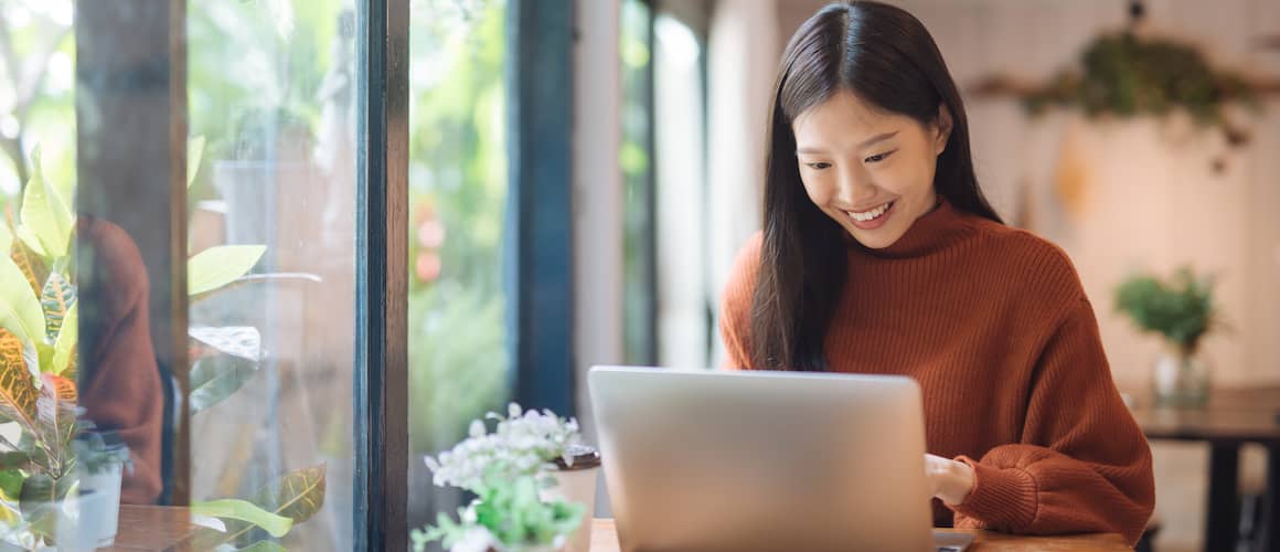 Smiling Asian woman in clay colored sweater working on laptop next to windows.