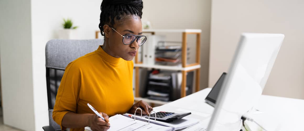 Woman with yellow shirt on computer with calculator doing taxes.
