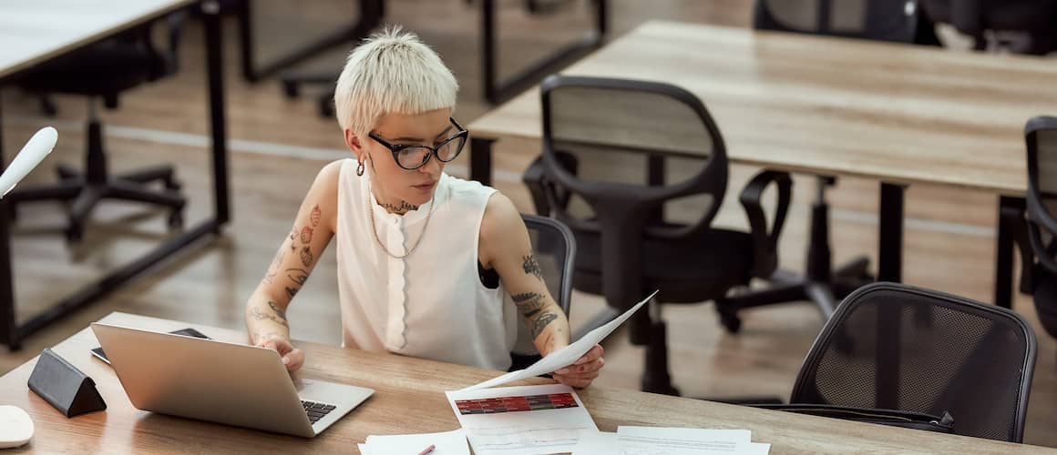 Young Woman With Tattoos And Short White Hair And Black Glasses Working On Laptop With Paperwork