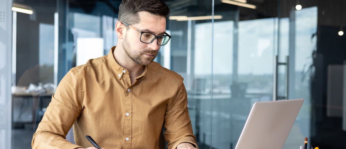 young man working at laptop while marking printed spreadsheet