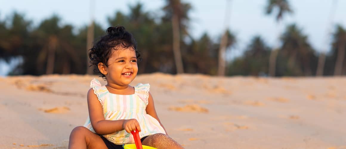 Toddler-age girl with curly black hair playing with bucket and shovel on beach and laughing.