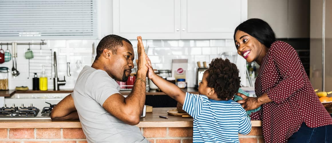 Father giving young son high five and mother smiles nearby all on kitchen stools.
