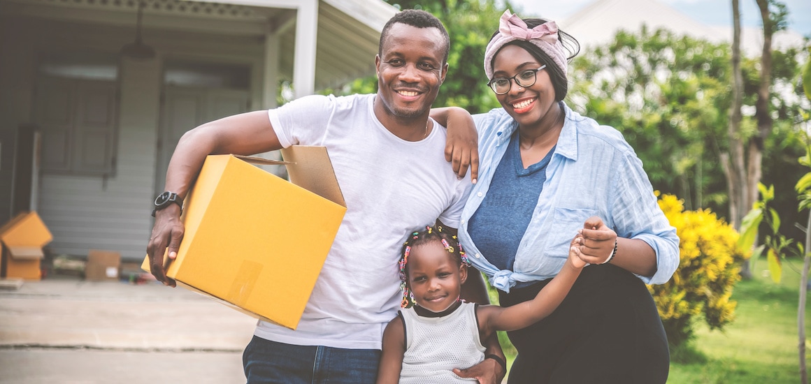 Man, woman and girl standing outside home holding a moving box and smiling.