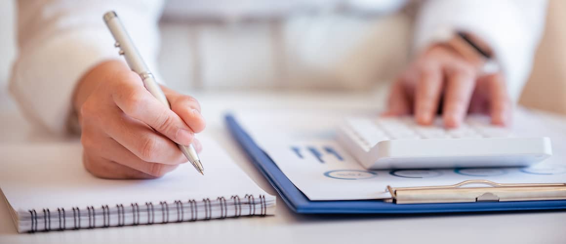 One hand pressing on a calculator while the other hand holds a silver pen and writes on a notepad