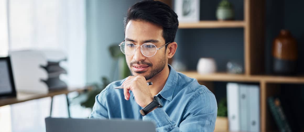 Business man with dark hair, wearing glasses and blue shirt, holding pen and looking at computer screen thinking.