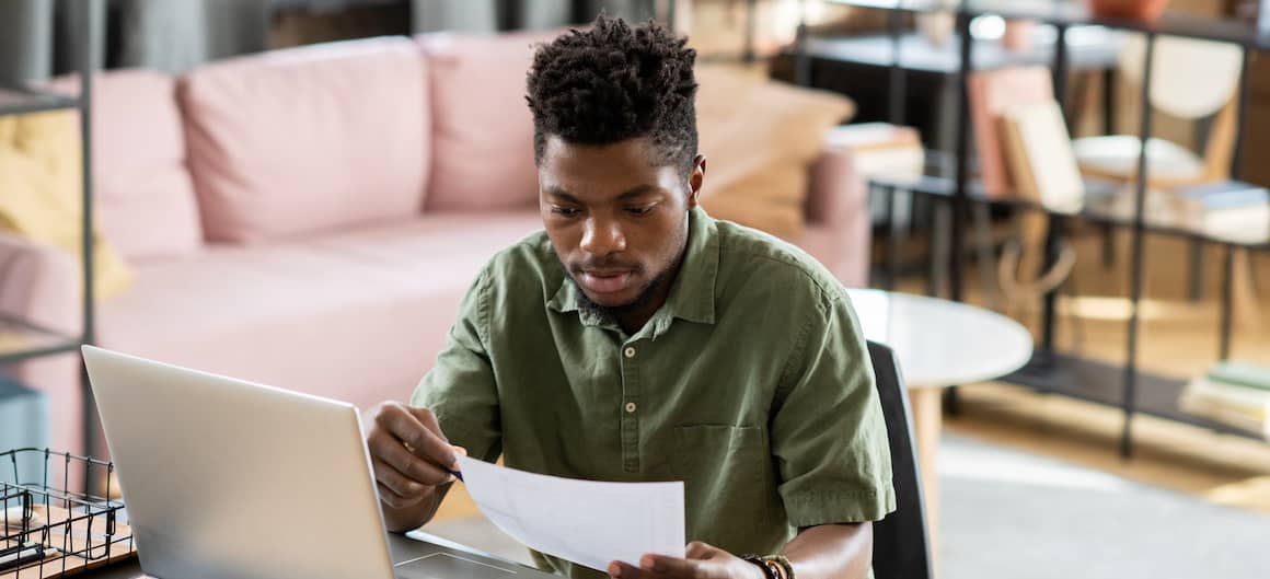 A young man working on paperwork and his laptop.