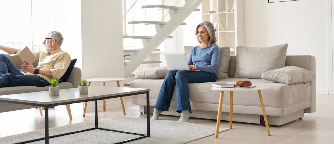 Senior couple relaxing on couches in living room while man reads a book and woman works on the computer.