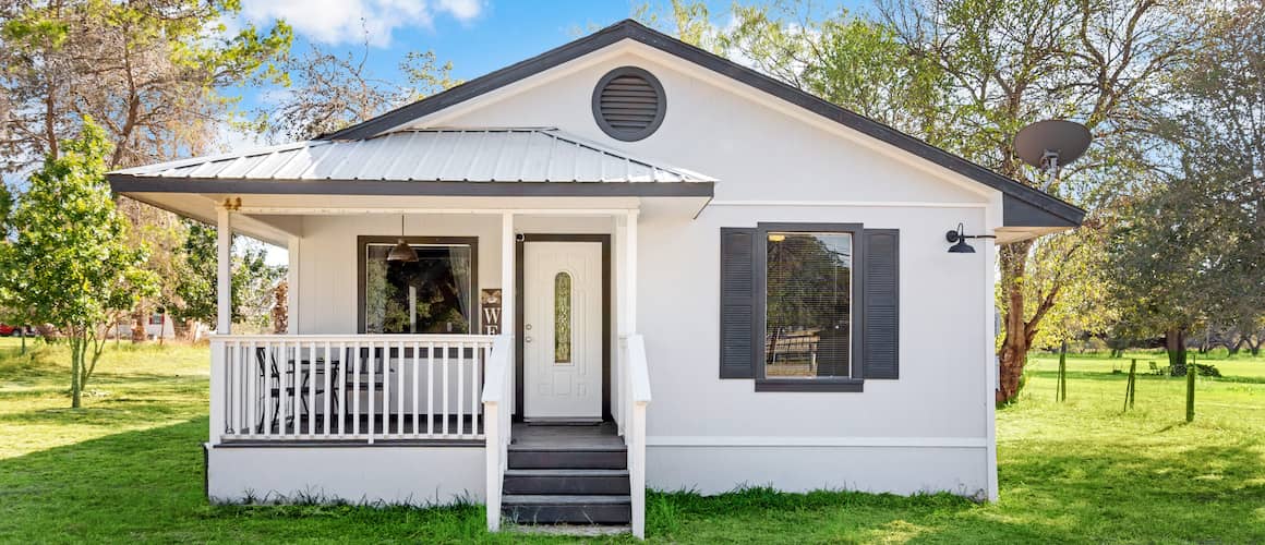 Small white, one-story house with gray trim, two windows, porch and metal roof.