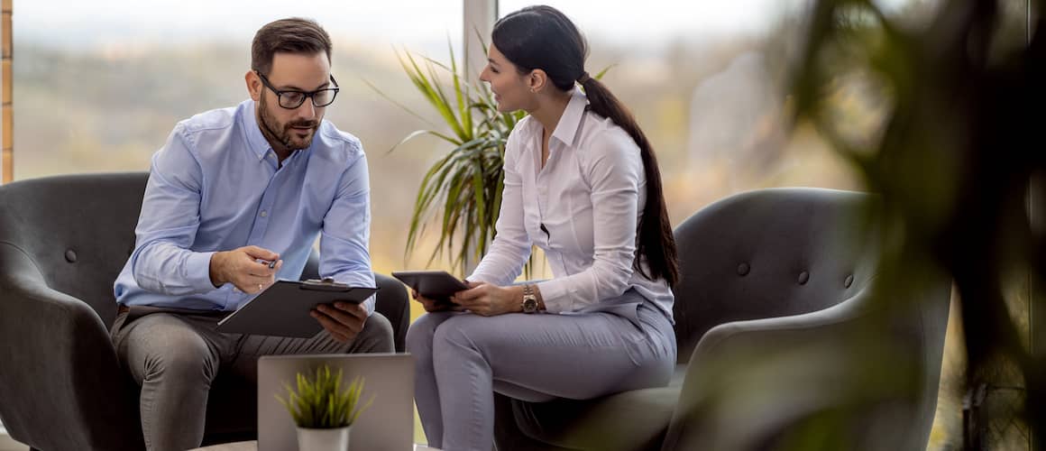 Two people in business attire talking together while sitting down on armchairs.