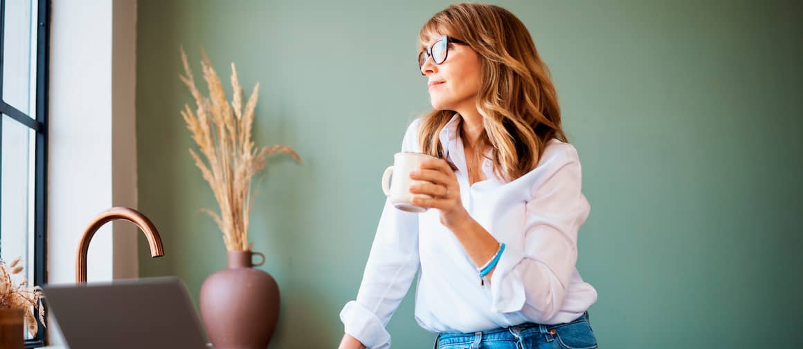Woman standing in green kitchen with laptop open, drinking coffee while looking out the window.