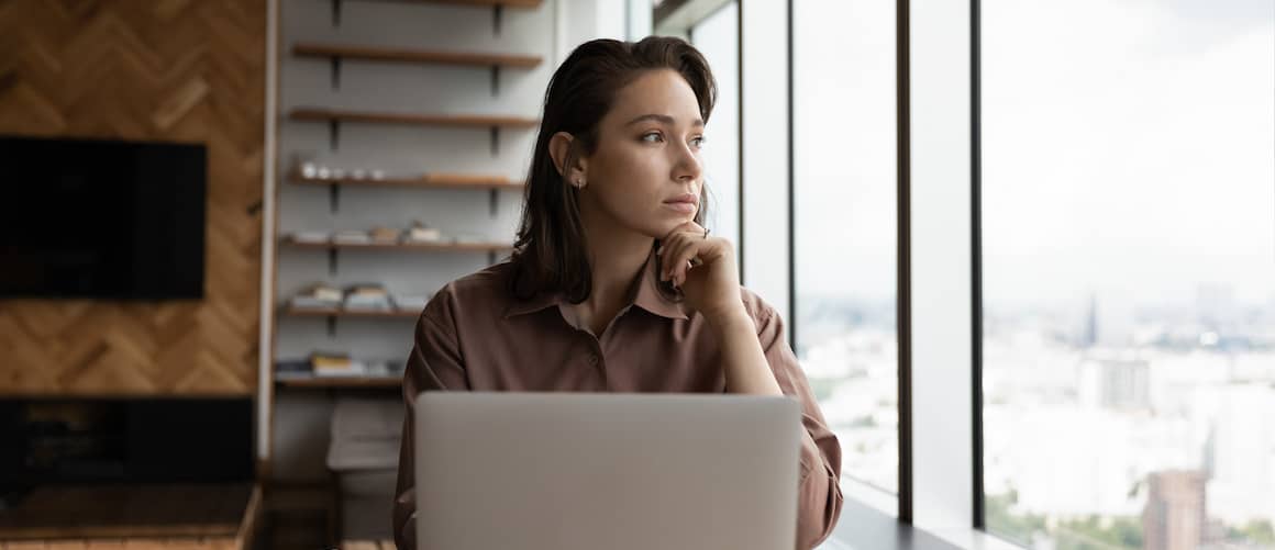 Young woman sitting at table with computer and documents looking out window, thinking.