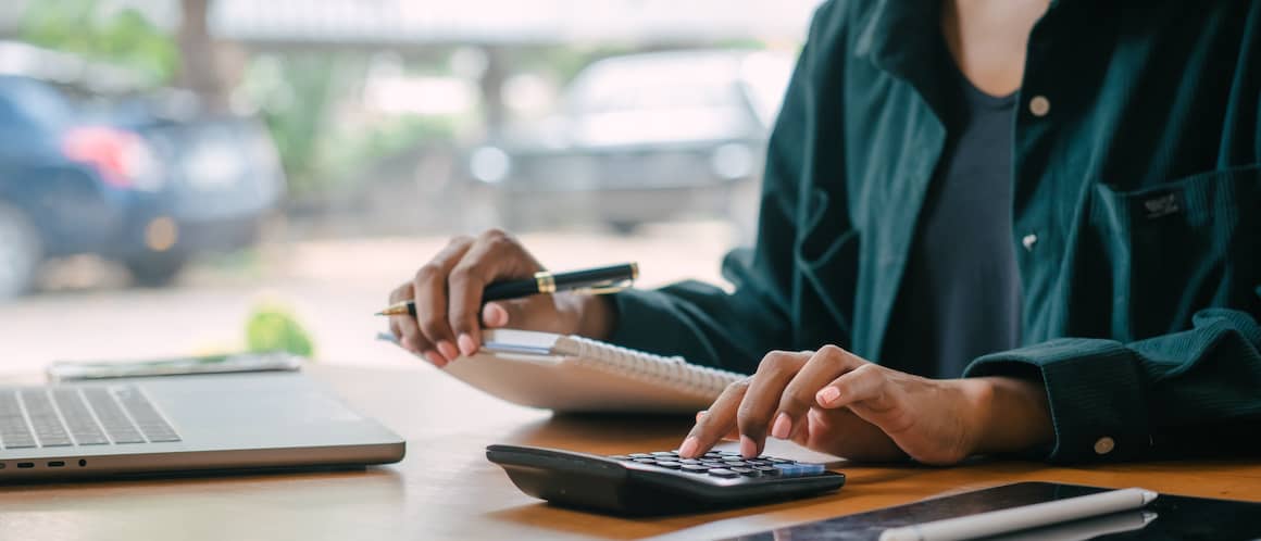 Close up of person sitting at desk, holding notebook and pen and using a calculator.