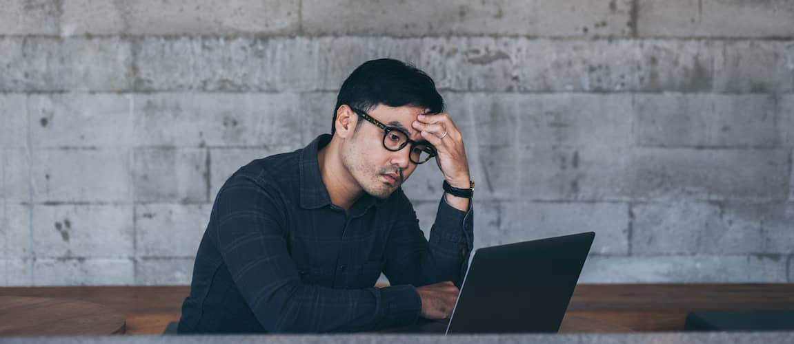 Young Asian man looking at his laptop seemingly stressed out in front of a gray brick wall.
